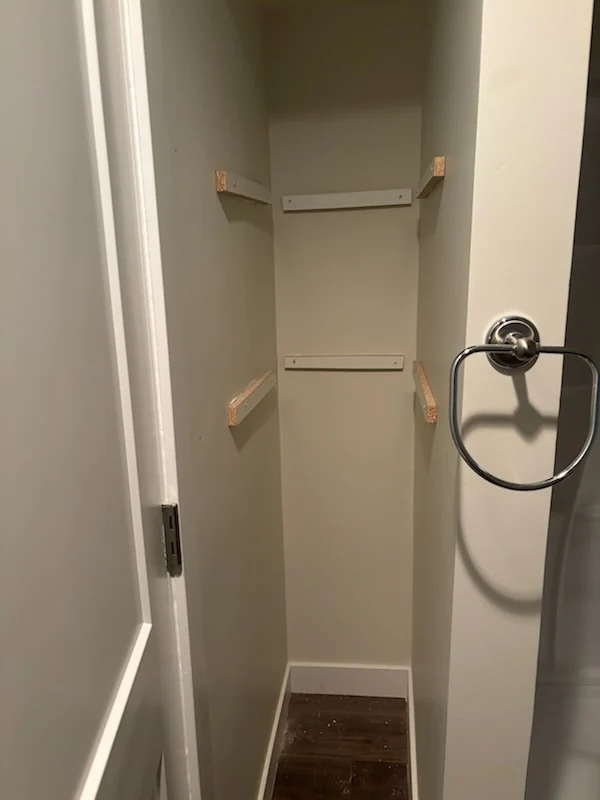 Small white closet with three installed shelves above a dark wood-look floor.