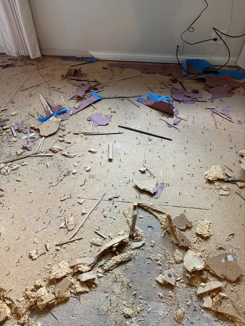 Room floor during covering removal, with torn underlay, strips, and loose debris beside a white wall.