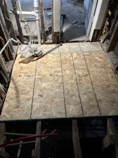 Wood subfloor panels laid across part of an opened floor, with exposed joists in the foreground.