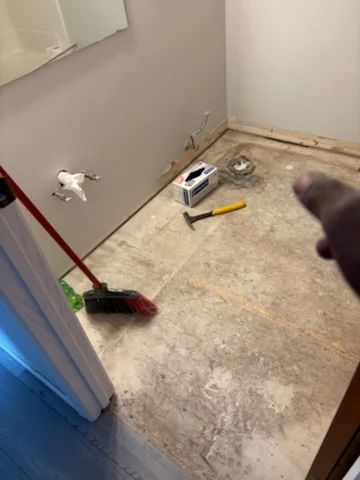 Bathroom floor during cleanup with broom, tools, and loose debris visible.