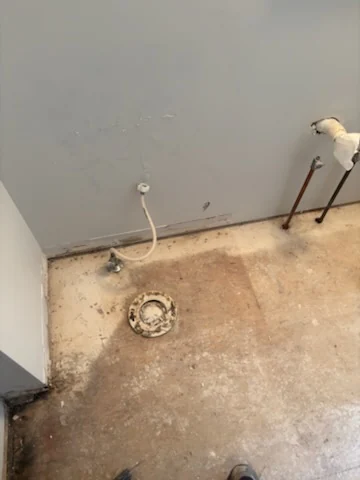 Bathroom floor after tile removal with the toilet removed and plumbing connections exposed.