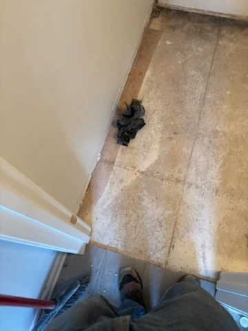 Bathroom doorway during floor tile removal with stripped flooring and work shoes visible.