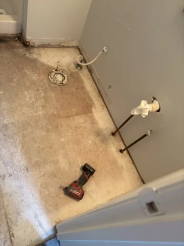 Small bathroom floor after tile removal with a drill and exposed toilet connection.