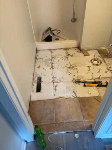 Bathroom floor during tile removal near a shower entry, with tools and remaining tile visible.